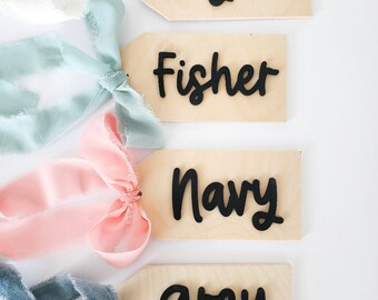 Può includere: Quattro etichette regalo in legno con nomi scritti in corsivo nero. I nomi sono Cayden, Fisher, Navy e Grey. Ogni etichetta è legata con un nastro di colore diverso.