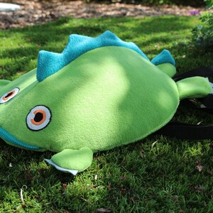 May include: A green plush backpack shaped like a dinosaur with blue spikes and orange eyes. The backpack has a zipper closure and a black adjustable strap.