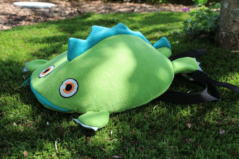 May include: A green plush backpack shaped like a dinosaur with blue spikes and orange eyes. The backpack has a zipper closure and a black adjustable strap.