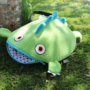 May include: Green plush monster backpack with a zippered mouth, large orange eyes, and a blue and white patterned lining.