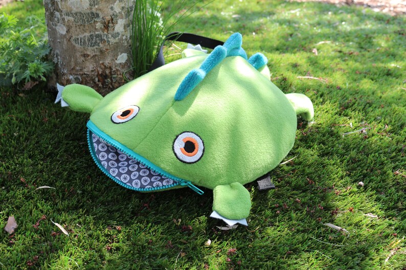 May include: Green plush monster backpack with a zippered mouth, large orange eyes, and a blue and white patterned lining.