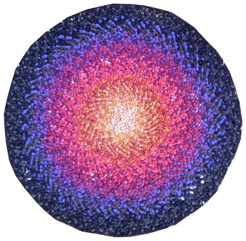Peut inclure: Un tapis rond avec un motif en spirale. Le centre est blanc chaud qui se fond dans des nuances de jaune, de rose et de violet. Le bord ext&eacute;rieur est bleu fonc&eacute;. Le tapis est fait de petits morceaux textur&eacute;s.