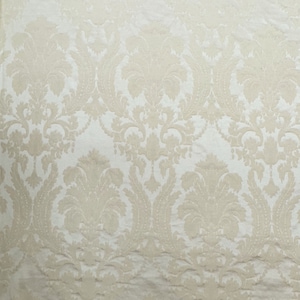 May include: Cream-colored fabric with a repeating damask pattern. The design features large, symmetrical floral and leaf motifs in a raised, textured style against a lighter background. The fabric has a subtle sheen.