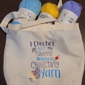 Puede incluir: Una bolsa de lona blanca con un diseño colorido que dice "Yo tejo pero mi pasatiempo favorito es coleccionar hilo". La bolsa está llena de tres ovillos de hilo azul, amarillo y morado.