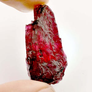 May include: A deep red, faceted, rough-cut crystal with a pointed tip. The crystal has a translucent appearance and is irregularly shaped.