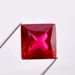 May include: A vibrant red square-shaped gemstone with a faceted cut. The gemstone is held by a pair of tweezers.