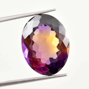 Pode incluir: Uma pedra preciosa de corte oval, segurada por uma pinça. A gema apresenta um gradiente de cores, incluindo roxo, amarelo e laranja. As facetas da pedra refletem a luz, criando um efeito brilhante. A gema está sobre um fundo branco.