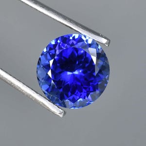 May include: A round, deep blue gemstone with a faceted cut. The gemstone is held by a pair of tweezers.