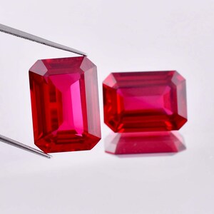 May include: Two vibrant red emerald-cut gemstones, likely rubies, are displayed on a white surface. One gemstone is held by tweezers, showcasing its brilliance and clarity.