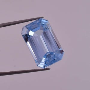 May include: A light blue gemstone with an emerald cut. The gemstone is held by tweezers.