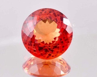 May include: A faceted, round gemstone with a vibrant orange-red hue. The gem's surface reflects light, creating a sparkling effect. The gemstone is displayed on a reflective surface, showing its mirrored image. This gemstone is a beautiful example of fine jewelry.