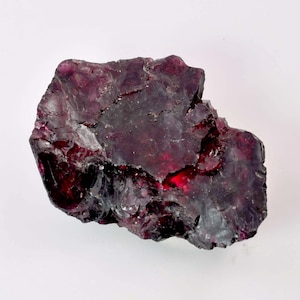 May include: A rough, irregularly shaped, dark red-purple gemstone with a crystalline structure. The stone has a deep, rich color with areas of lighter red and a slightly reflective surface. The gemstone is set against a white background.