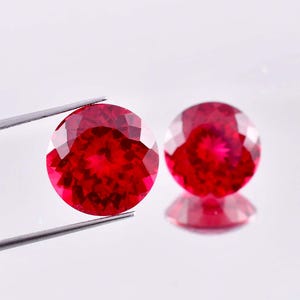 Könnte beinhalten: Zwei runde, rote Edelsteine mit facettiertem Schliff. Die Edelsteine liegen auf einem weißen Hintergrund.