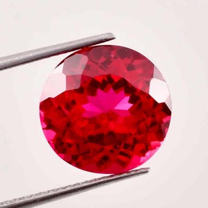 Puede incluir: Una gema roja vibrante con un corte redondo y una superficie facetada. La gema se sostiene con unas pinzas sobre un fondo blanco.