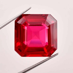 May include: A vibrant red gemstone with a square emerald cut. The gemstone is held by tweezers against a white background.