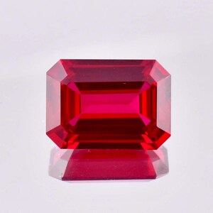 May include: A deep red emerald cut gemstone with a high polish. The gemstone is reflecting light and has a slight shadow underneath.