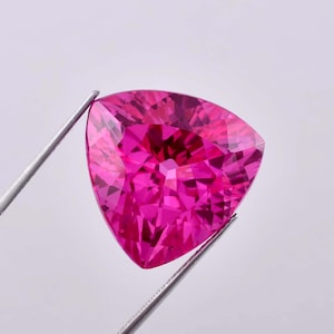 May include: A vibrant pink gemstone with a triangular cut. The gemstone is held by a pair of tweezers.