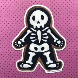 May include: A brown and white skeleton illustration of a person wearing a black hooded sweatshirt. The illustration is on a pink background with a white pattern.
