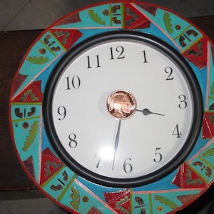 May include: A round wall clock with a white face and black hands. The clock's bezel is decorated with a colorful geometric pattern in red, turquoise, and blue. A copper coin is in the center of the clock face.