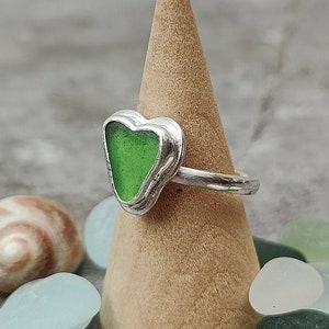 Op de afbeelding: Een zilveren ring met een groen, hartvormig stuk zeeglas. De ring wordt gepresenteerd op een houten kegel.