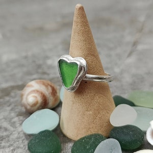 Op de afbeelding: Een zilveren ring met een groen, hartvormig stuk zeeglas. De ring wordt getoond op een houten ringstandaard, omringd door zeeglas en schelpen.