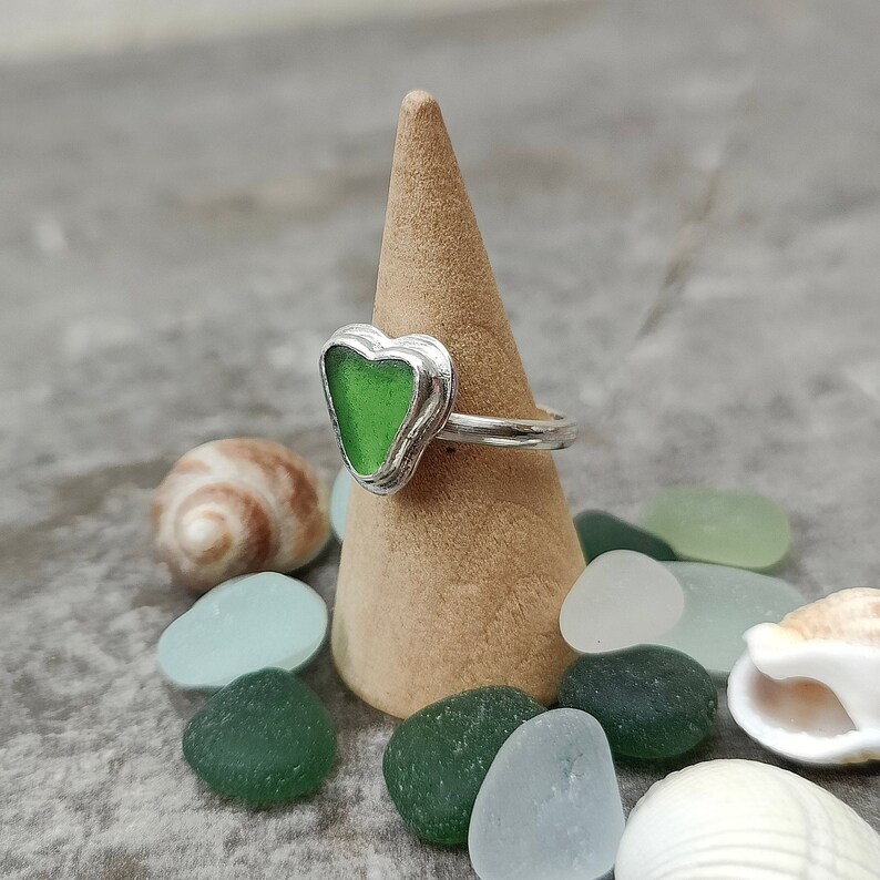 Op de afbeelding: Een zilveren ring met een groen, hartvormig stuk zeeglas. De ring wordt getoond op een houten ringstandaard, omringd door zeeglas en schelpen.