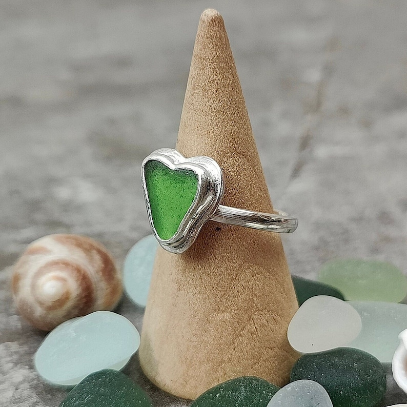 Op de afbeelding: Een zilveren ring met een groen, hartvormig stuk zeeglas. De ring wordt gepresenteerd op een houten kegel.