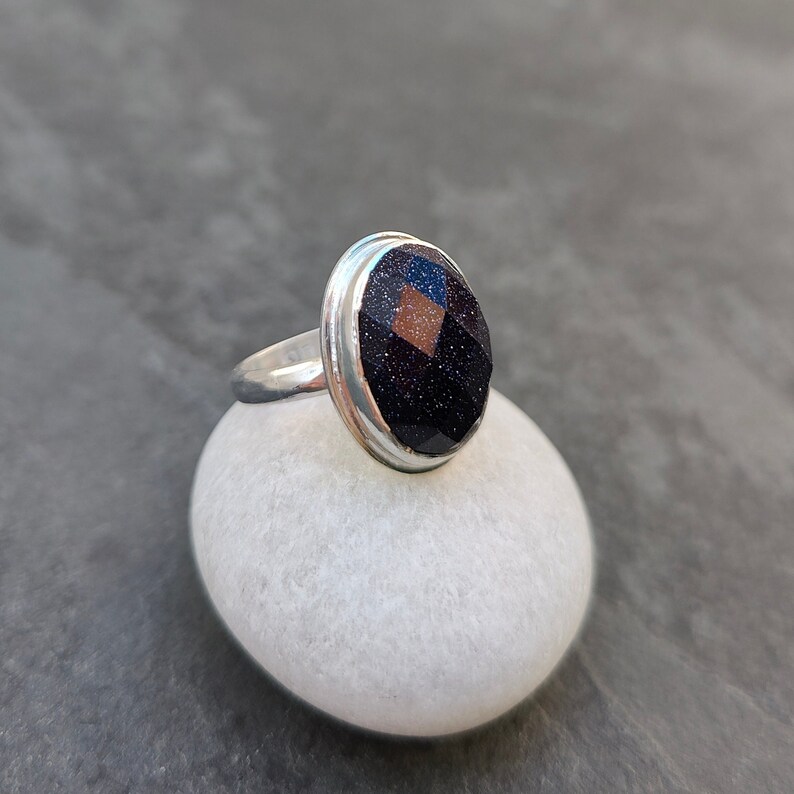 Puede incluir: Un anillo de plata con una piedra ovalada de color azul oscuro. La piedra tiene una textura brillante y reluciente.