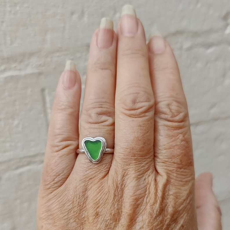 Op de afbeelding: Een zilveren ring met een groen, hartvormig stuk zeeglas dat in het midden is gezet.