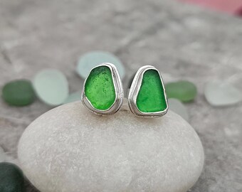 Op de afbeelding: Een paar zilveren oorbellen met groen zeeglas in het midden. De oorbellen liggen op een wit stenen oppervlak.