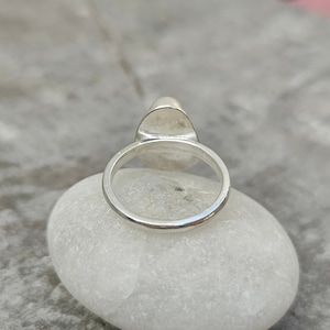 May include: A silver ring with a smooth, oval-shaped stone set in the center. The ring is sitting on a white rock.