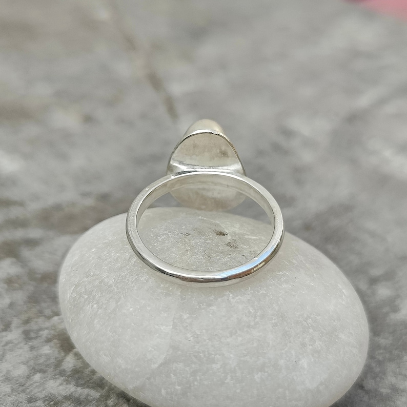 May include: A silver ring with a smooth, oval-shaped stone set in the center. The ring is sitting on a white rock.