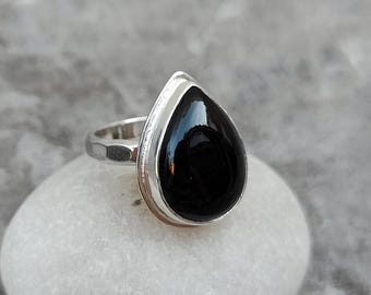 以下が含まれることがあります： 大きな涙型の黒い宝石が付いたシルバーリング。磨かれた宝石は光を反射します。リングバンドはテクスチャード加工のデザインです。リングは、ぼやけた灰色の背景に対して、白く丸みを帯びた表面にセットされています。