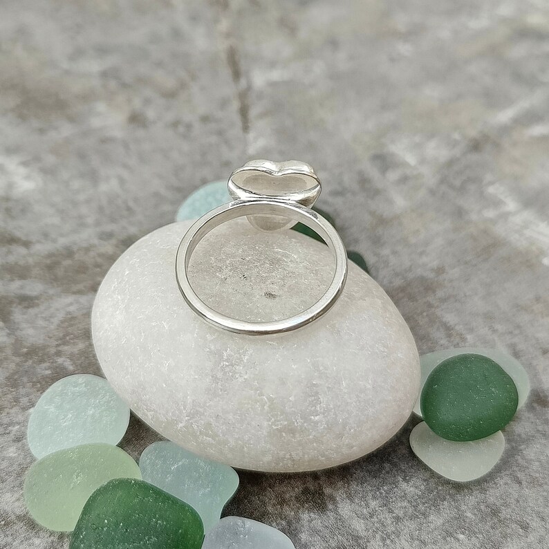 Op de afbeelding: Een zilveren ring met een hartvormig ontwerp. De ring ligt op een witte steen.