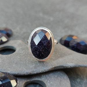 Puede incluir: Un anillo de plata con una piedra preciosa azul oscuro facetada. La piedra preciosa tiene una textura brillante y moteada.