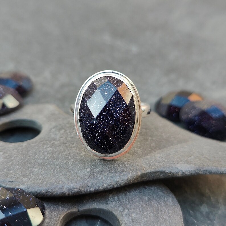 Puede incluir: Un anillo de plata con una piedra preciosa azul oscuro facetada. La piedra preciosa tiene una textura brillante y moteada.