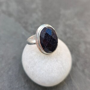 Puede incluir: Un anillo de plata con una piedra preciosa ovalada de color azul oscuro. La piedra preciosa tiene una superficie texturizada y brillante.