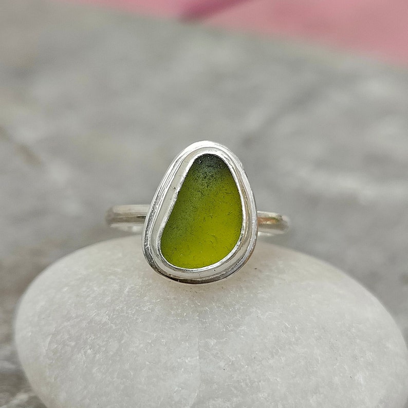 May include: A silver ring with a green sea glass stone set in the center. The ring is on a white rock.