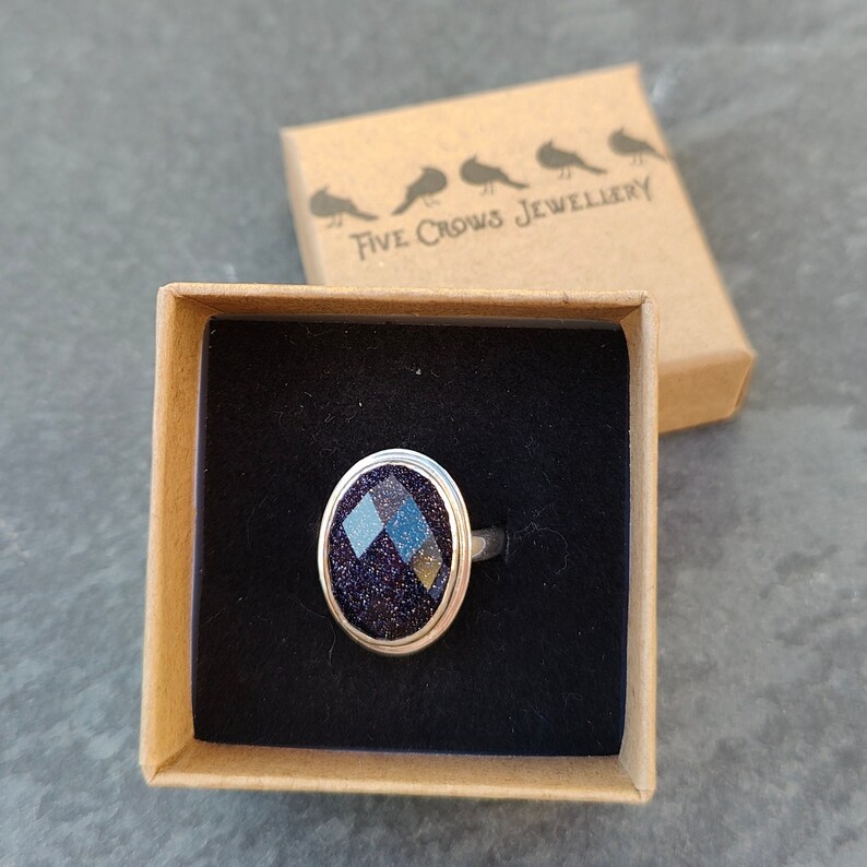 Puede incluir: Un anillo de plata con una piedra ovalada de color azul oscuro y brillante, engastada en un engaste de plata. El anillo est&aacute; en una peque&ntilde;a caja marr&oacute;n con el texto "Five Crows Jewellery" impreso en la tapa.