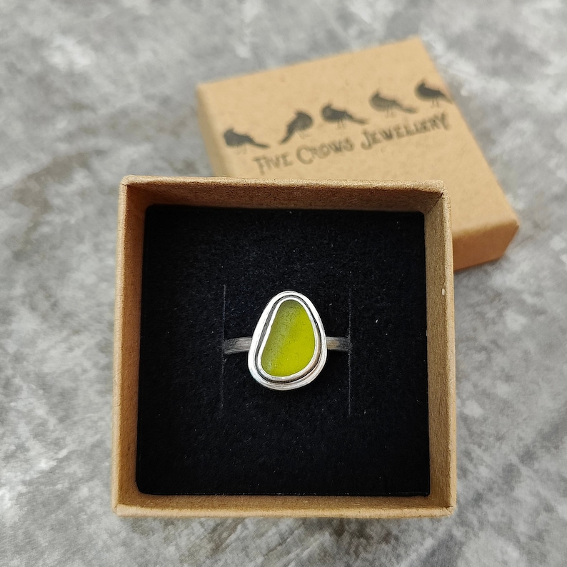 May include: A silver ring with a yellow sea glass stone set in a bezel setting. The ring is in a small brown box with the text "Five Crows Jewellery" on the lid.