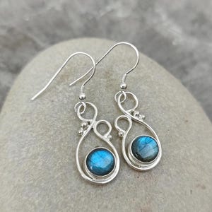 Puede incluir: Un par de pendientes colgantes de plata con piedras preciosas redondas de labradorita azul iridiscente engastadas en un marco de alambre metálico abierto y en espiral. Cada pendiente cuelga de un gancho de plata sencillo.