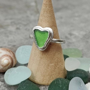 Op de afbeelding: Een zilveren ring met een groen, hartvormig stuk zeeglas dat in het midden is gezet. De ring wordt gepresenteerd op een houten ringstandaard.