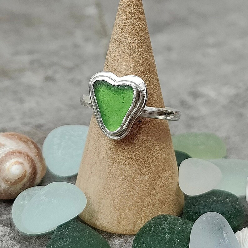 Op de afbeelding: Een zilveren ring met een groen, hartvormig stuk zeeglas dat in het midden is gezet. De ring wordt gepresenteerd op een houten ringstandaard.