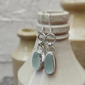 Puede incluir: Un par de pendientes colgantes con piedras ovaladas de color verde espuma de mar pálido y lisas, engastadas en plata. Cada pendiente cuelga de un gancho de plata unido a un anillo texturizado, conectado a un anillo menor que sujeta la piedra.