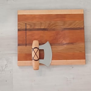 Op de afbeelding: Een houten snijplank met een decoratief houtnerfpatroon en een metalen kaassnijder met een houten handvat. Het mes heeft een gebogen lemmet en een decoratief ontwerp op het handvat.