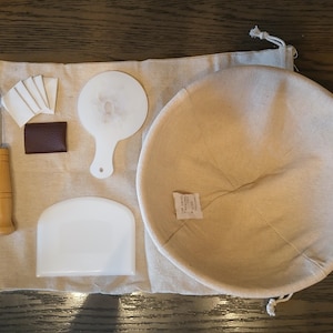 以下が含まれることがあります： パン作り用のベーキングツールセット。発酵バスケット、生地スクレーパー、スコアツール、ベンチスクレーパー、ラメラメが含まれています。ツールはベージュの布地に置かれています。