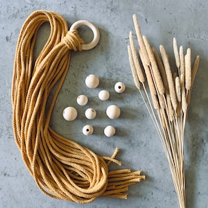 May include: A mustard yellow macrame cord bundle attached to a light-colored wooden ring, accompanied by nine small, round, unfinished wooden beads of varying sizes and a cluster of dried, tan-colored ornamental grass stalks.