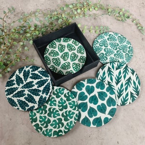 May include: A set of five circular coasters featuring various green leaf patterns on a cream background, stored in a black square holder. Each coaster displays a distinct botanical print, including monstera and heart-shaped foliage designs.