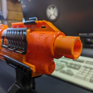 May include: Close-up of an orange and black toy gun with a black barrel and a black grip. The gun has a textured surface and a black and orange design.