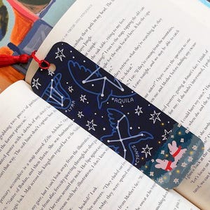 May include: Dark blue bookmark featuring constellations, including Lyra, Aquila, and Cygnus, and white stars. The bottom has a floral pattern and two pink rabbits. A red tassel hangs from the top. The bookmark is in an open book.